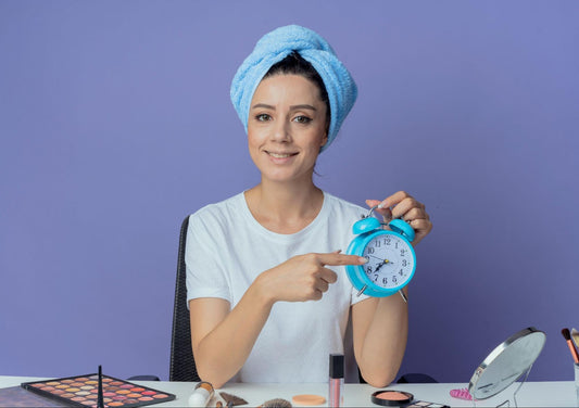 Woman with a towel on her head, holding an alarm clock, looking surprised in a bright, cozy room.