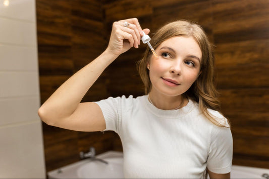 A woman is doing her skincare in a well-lit bathroom, standing in front of a mirror.