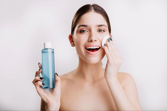 A woman holds a bottle of face wash in one hand and a bottle of water in the other, smiling at the camera.